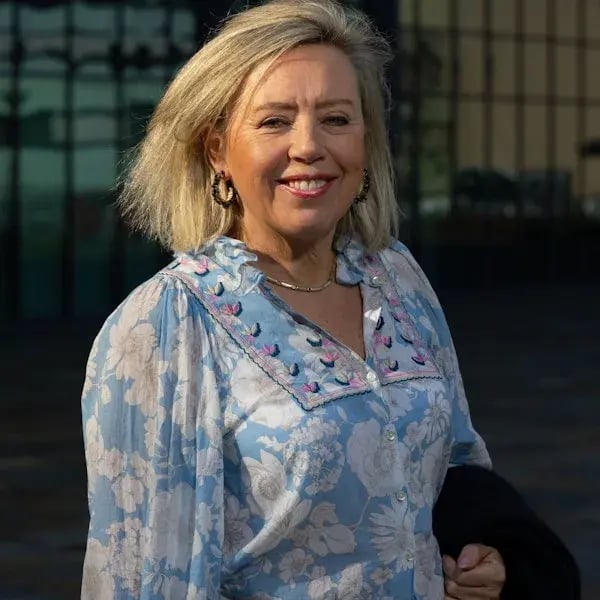 Een close-up van een vrouw in een bloemenblouse, lachend voor een glazen gebouw.