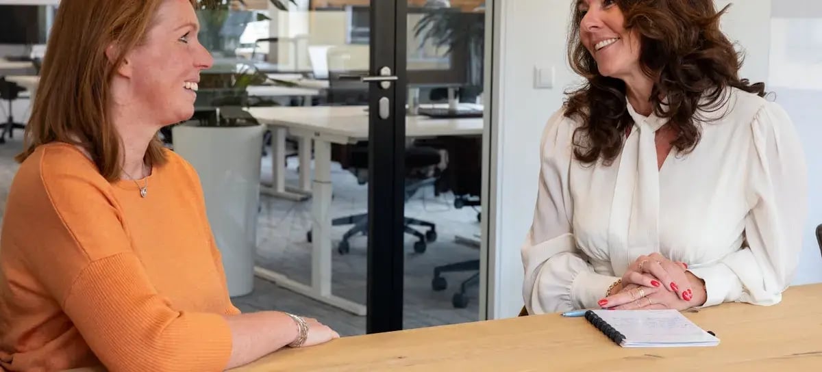 Twee vrouwen die aan een tafel in het kantoor zitten en met elkaar praten.