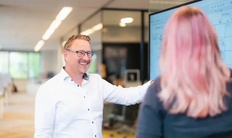 Man pointing at something on a large screen for a female colleague with pink hair.