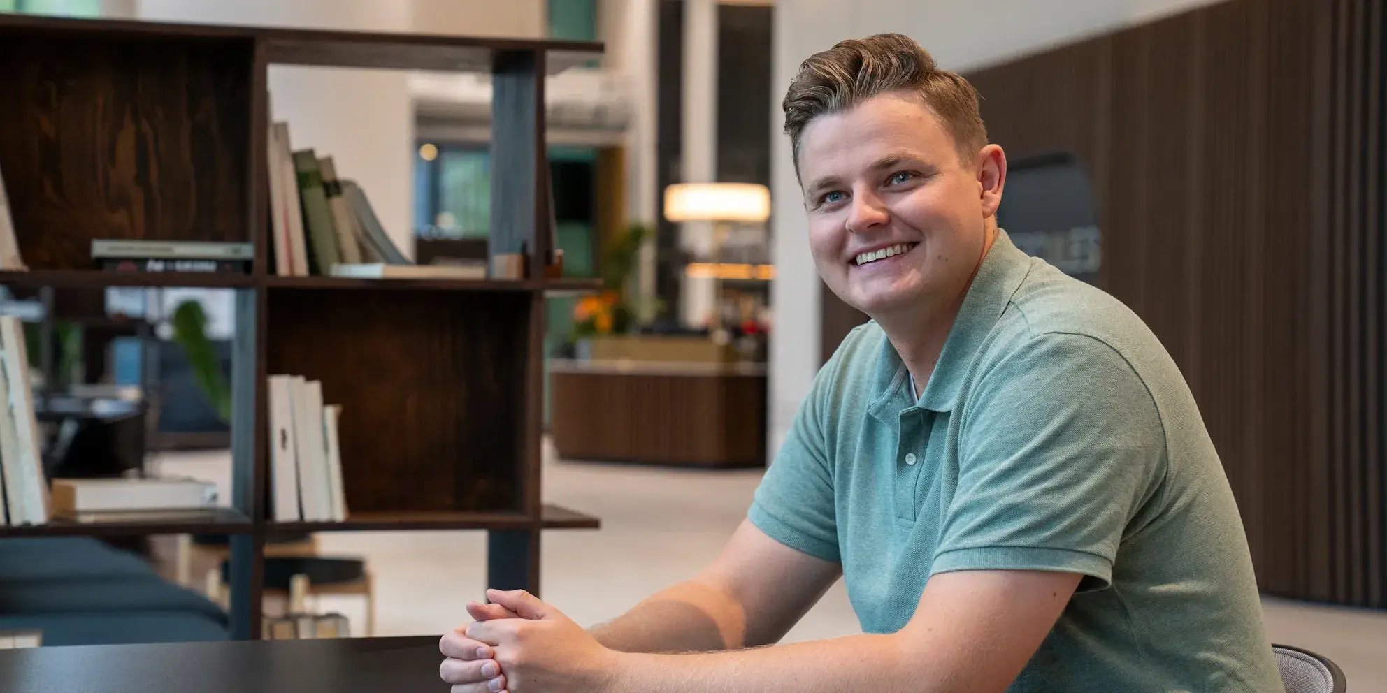 Jonge man in een groene polo poseert lachend aan een tafel in een kantoorruimte.
