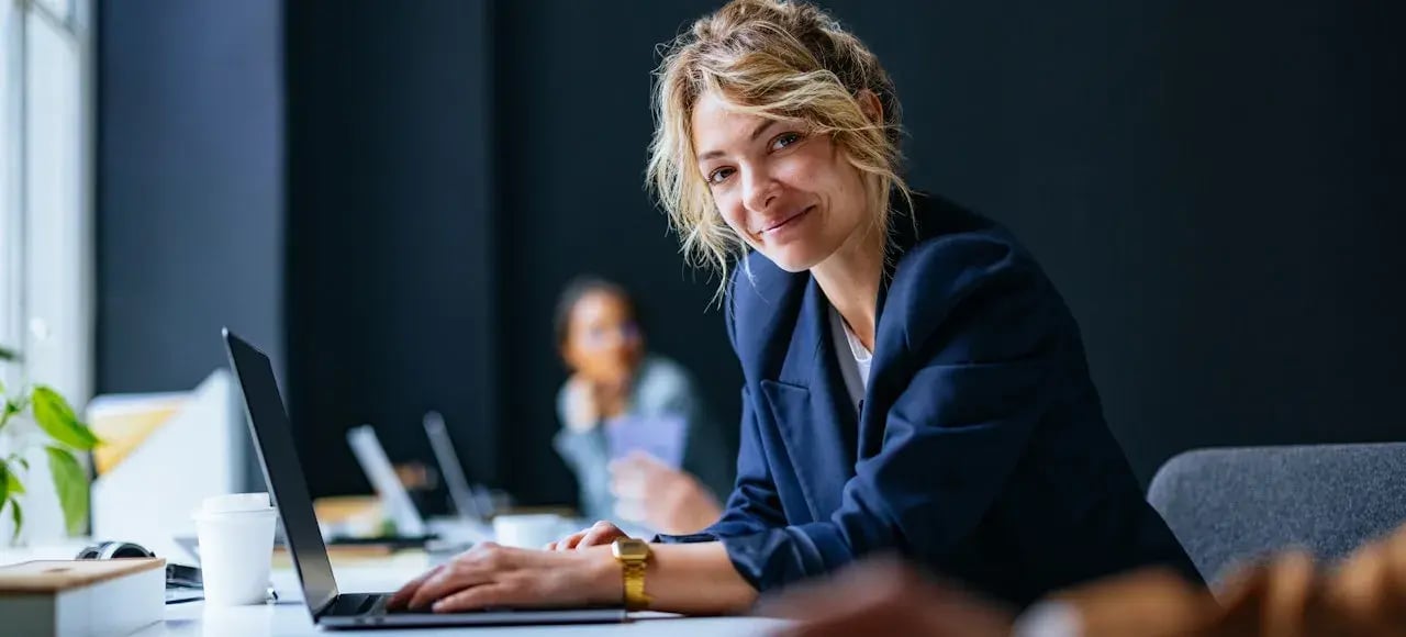 Woman with blonde hair behind her laptop, smiling at the camera.