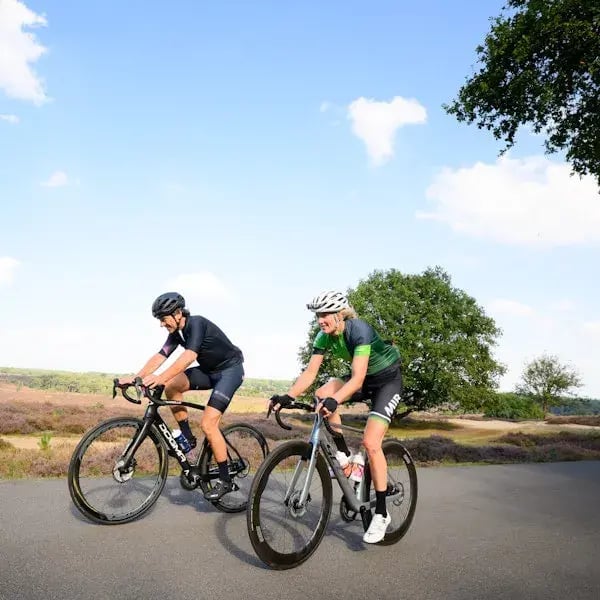 Twee wielrenners fietsen samen over een landelijke weg met heide en bomen op de achtergrond.