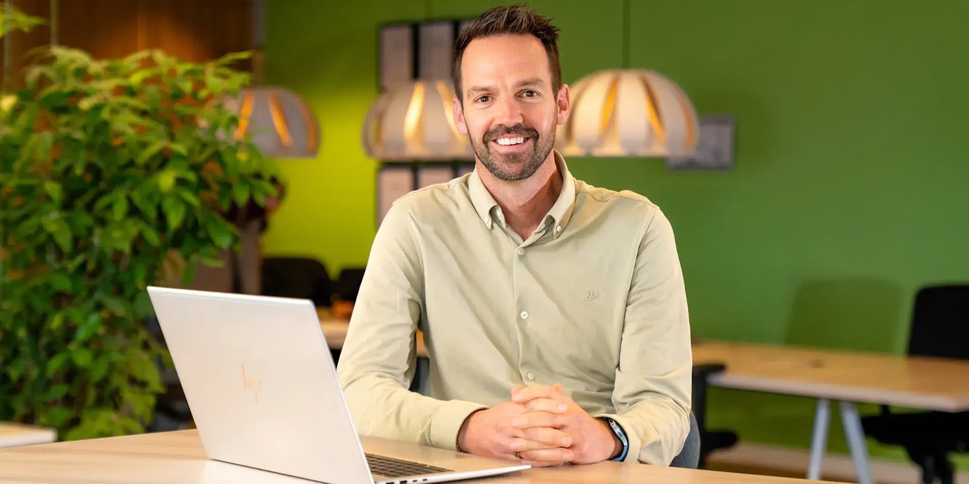 A man sits smiling behind a laptop in a modern office with a green wall.