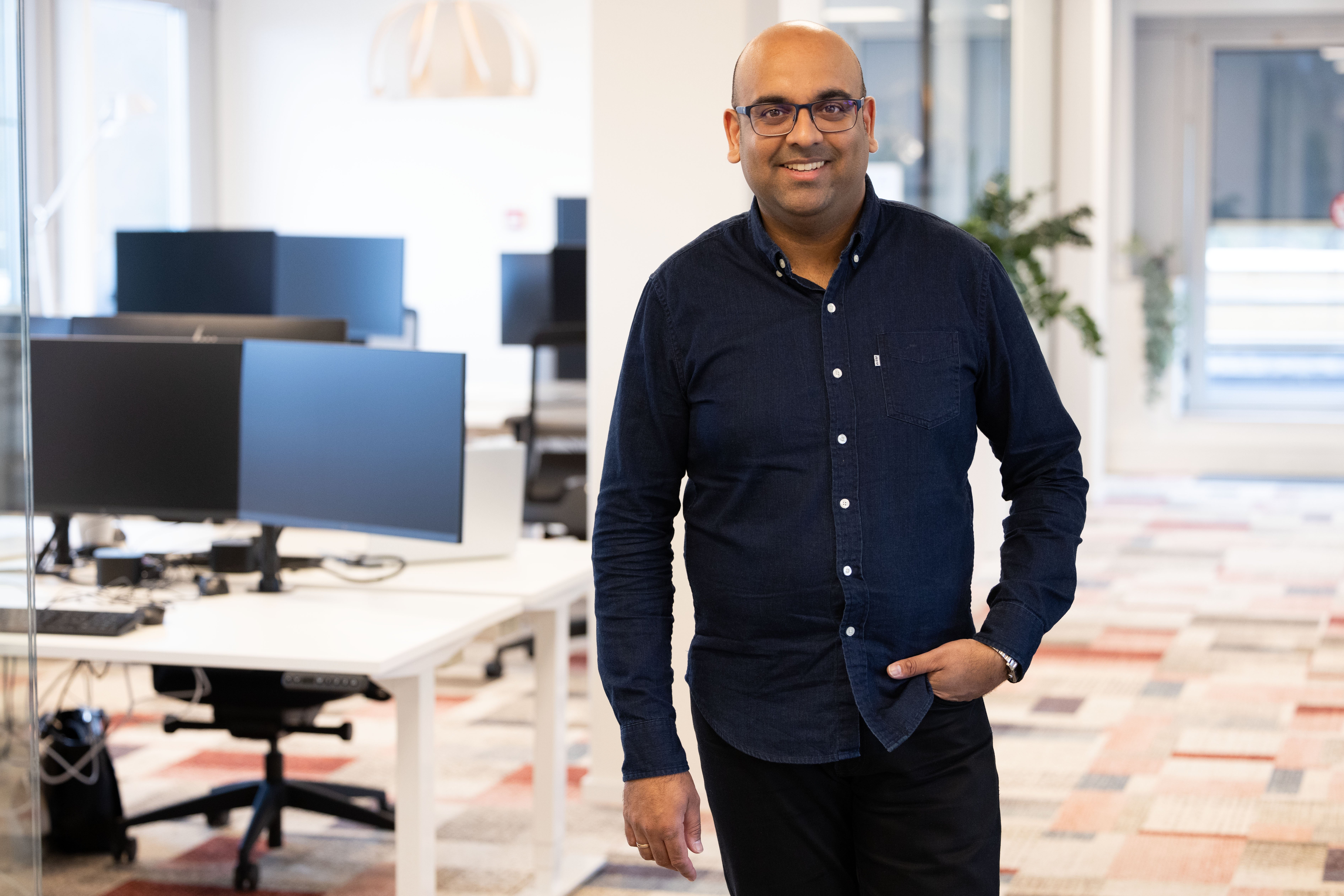 Man in a dark blue shirt stands in a modern office with desks and computers.