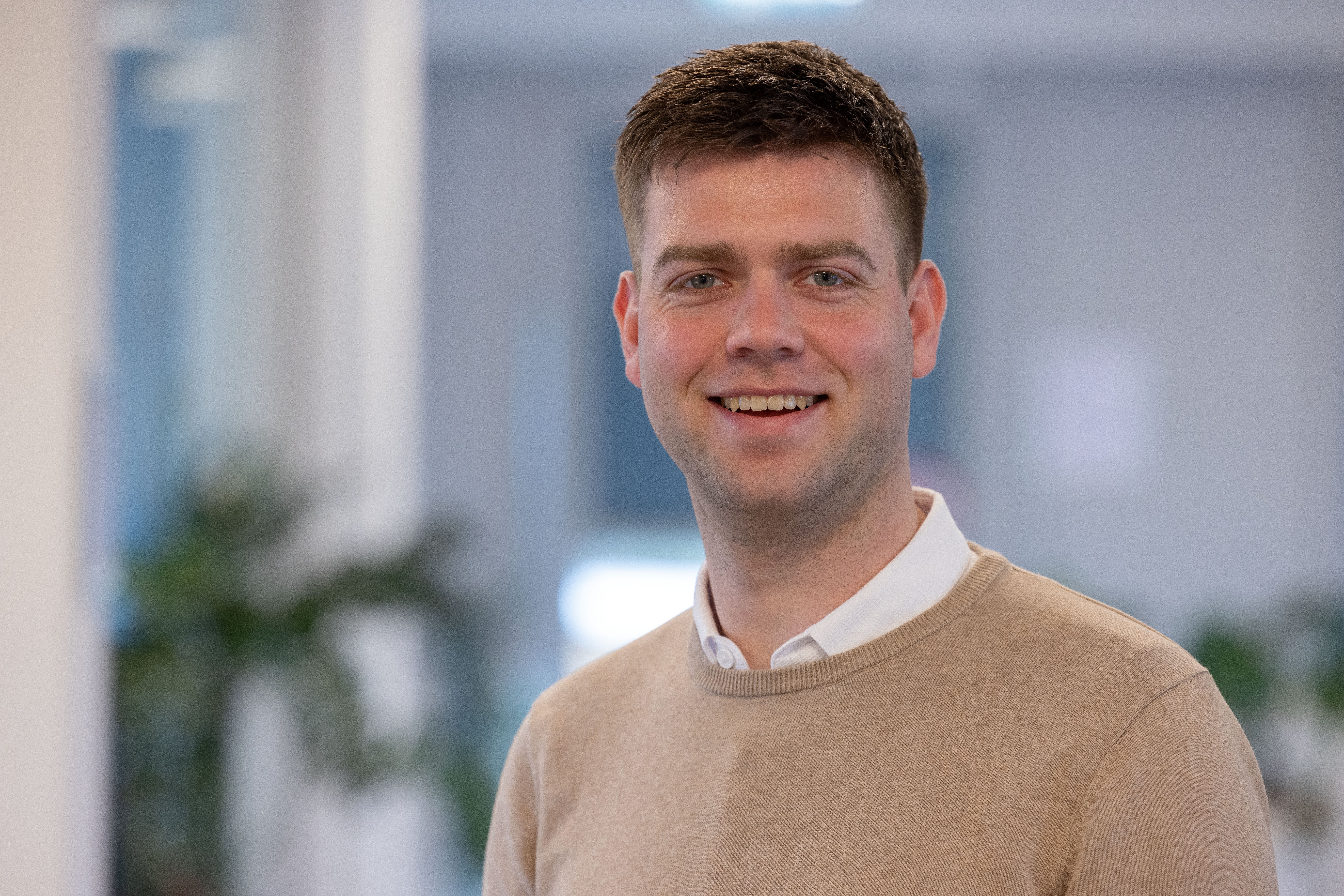 Man in beige sweater smiles at camera in an office.