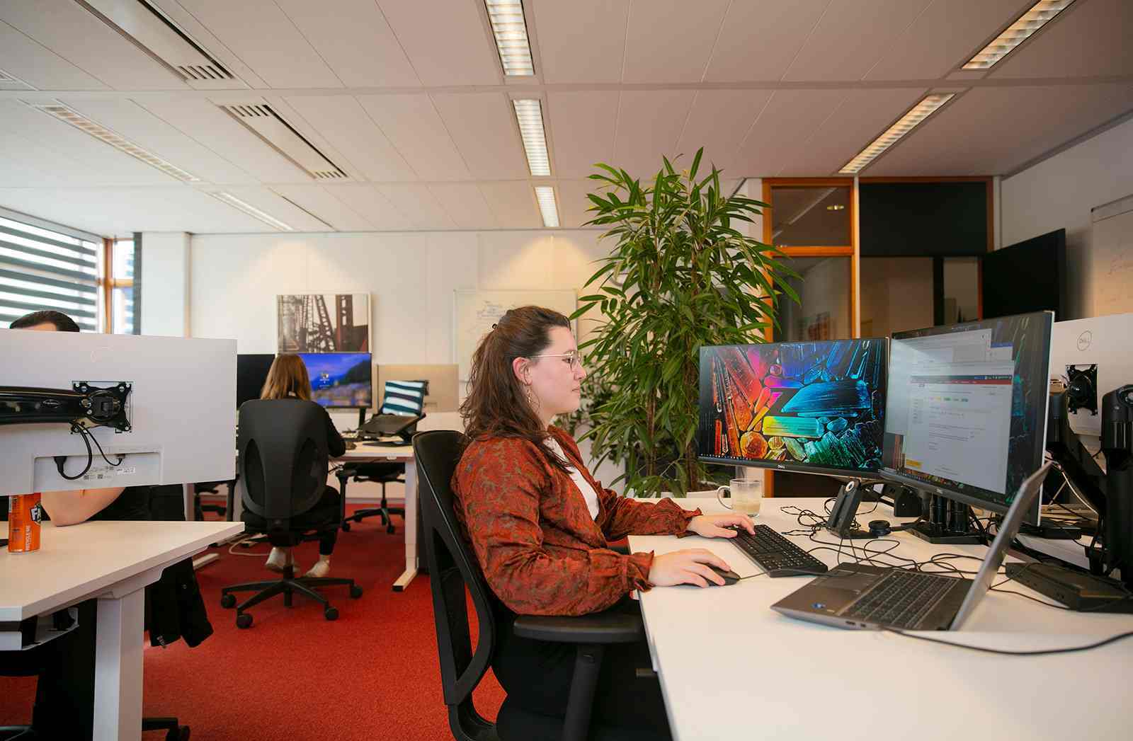 Vrouw werkt aan een bureau met twee beeldschermen in een kantoorruimte.