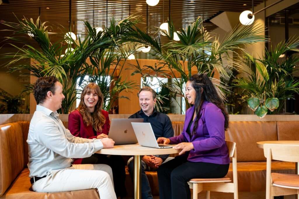 Group of four people laughing and working with laptops in an office environment.