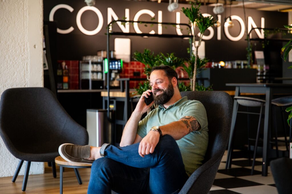 A man in a green shirt sits on a chair and talks on the phone in an office.