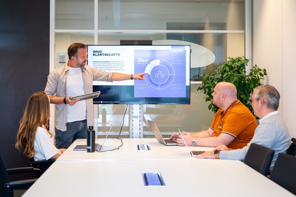 Man points to a screen, three people listen in a meeting room.
