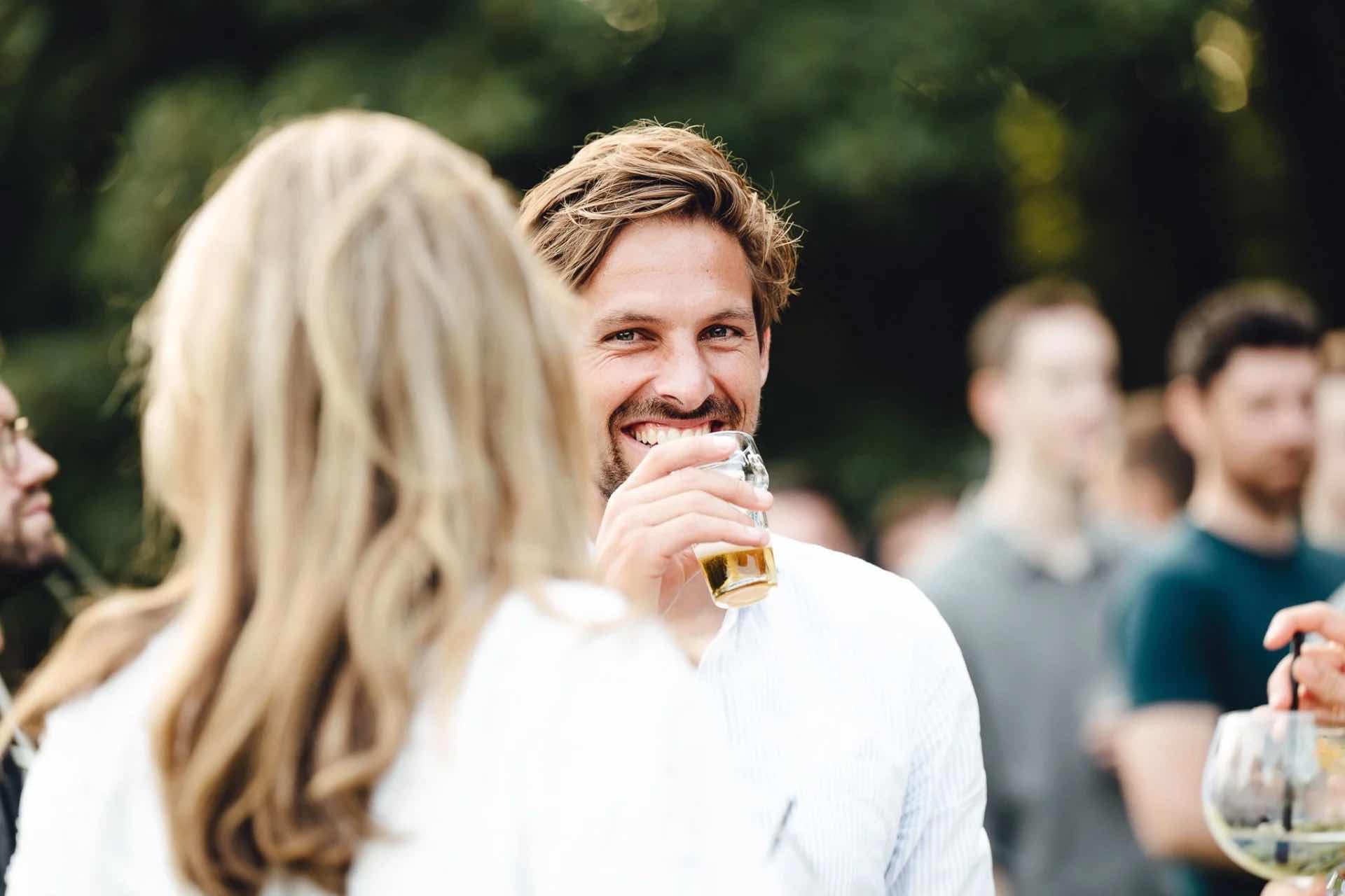 A man with a smile holds a glass of beer while talking to someone at an event.