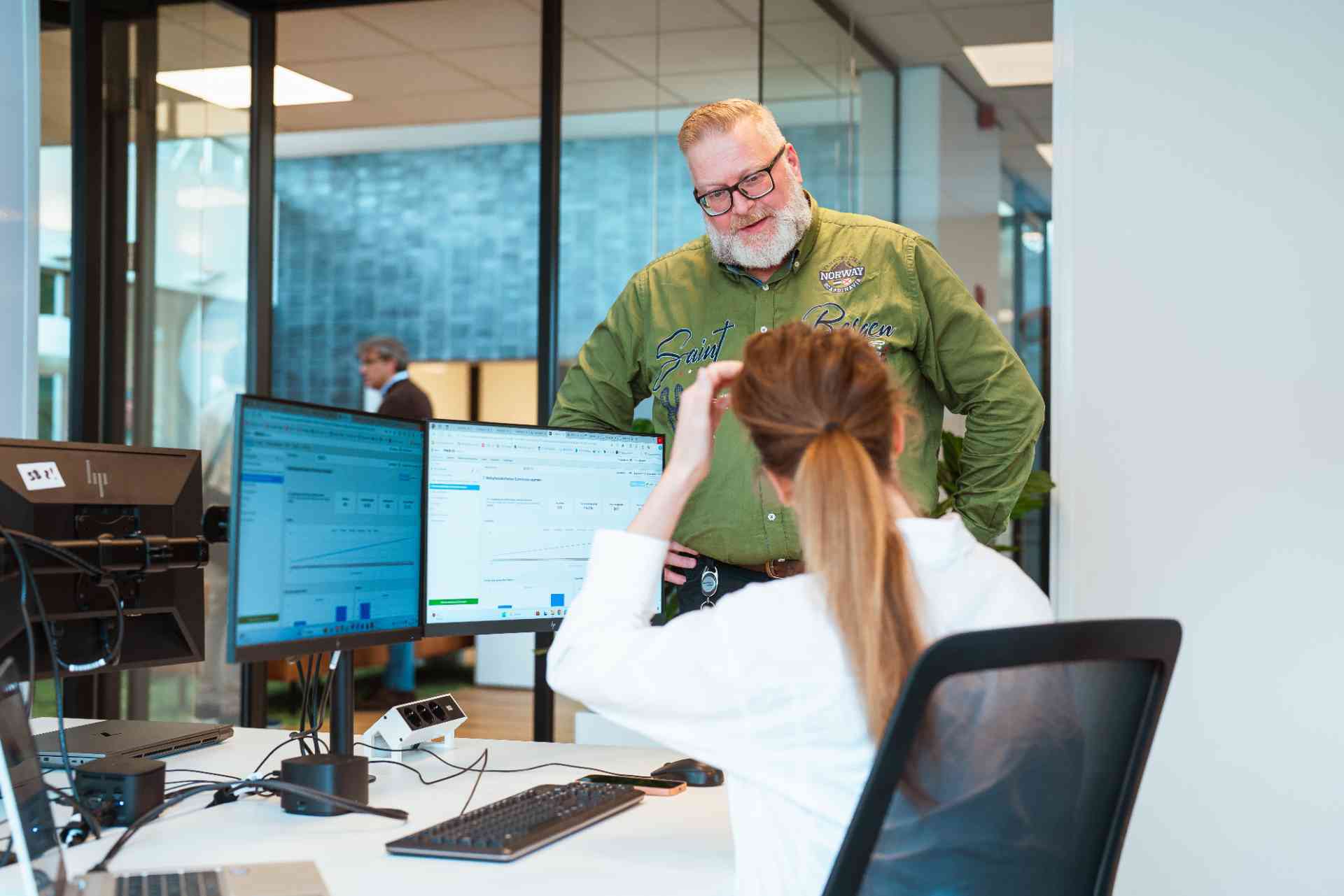 Twee mensen praten in een kantoor, terwijl een man naar een vrouw kijkt.
