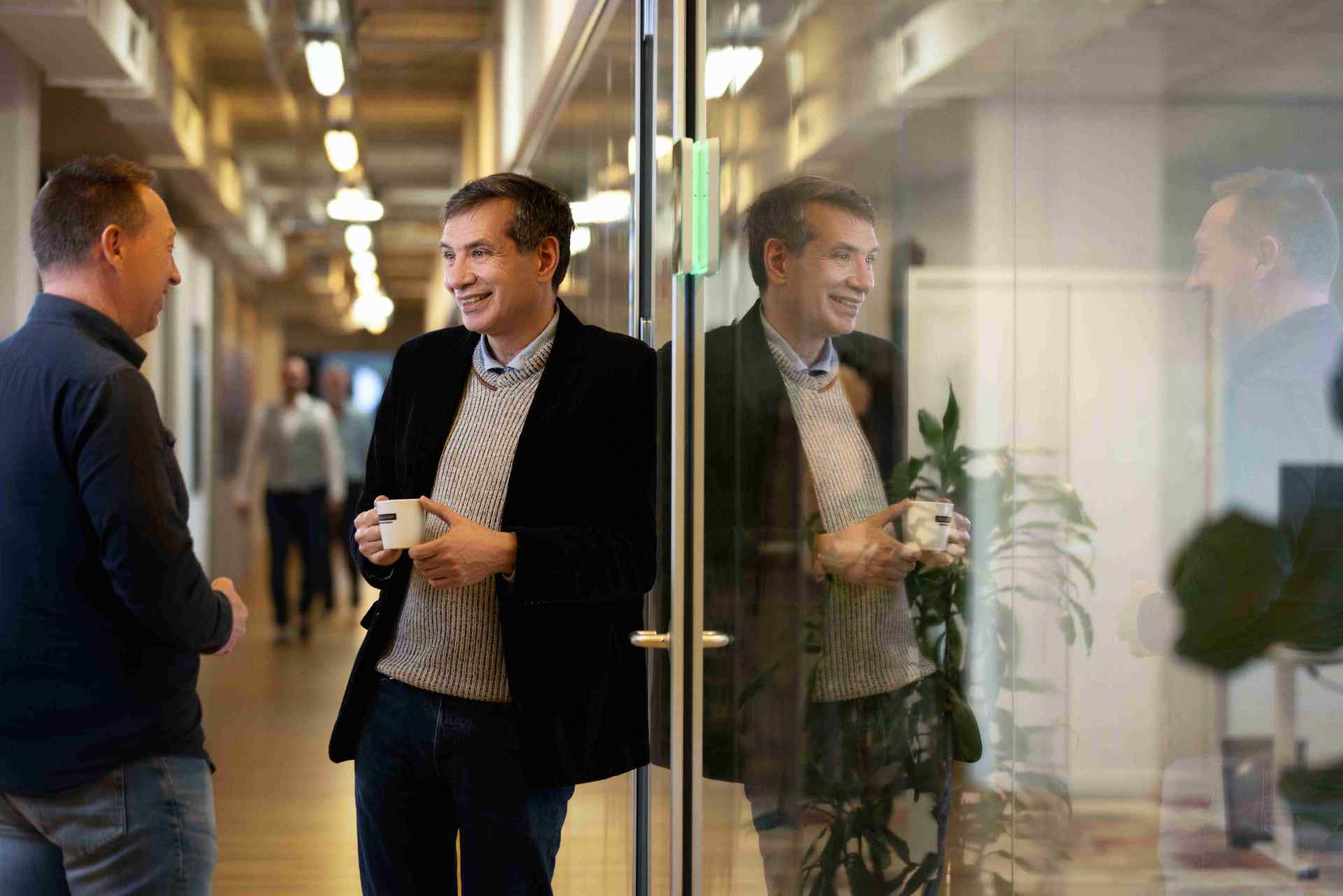 Two men talk in a hallway, while one man smiles with a mug in his hand.