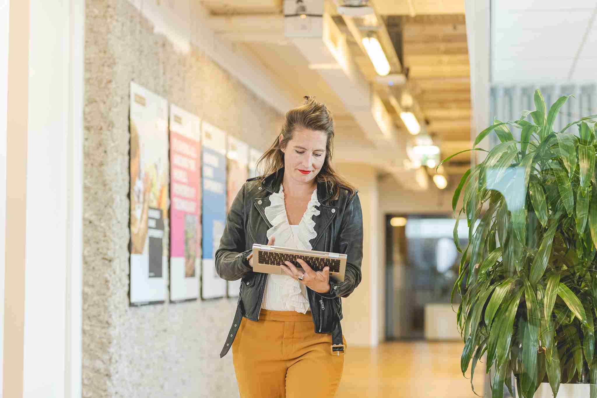 Woman in a leather jacket with a laptop in her hand walks through an office space.