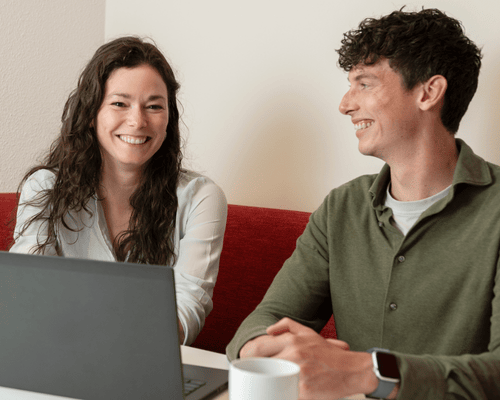 Two employees smile at each other as they sit at a table with a laptop and a cup in their hands.