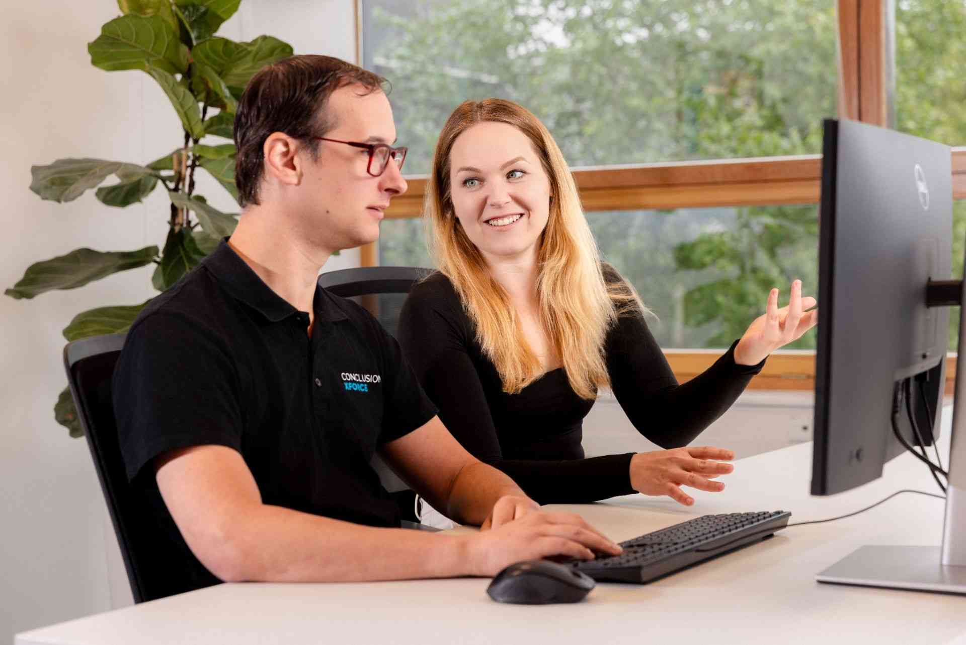 Two colleagues in an office laugh and talk while looking at a computer screen.