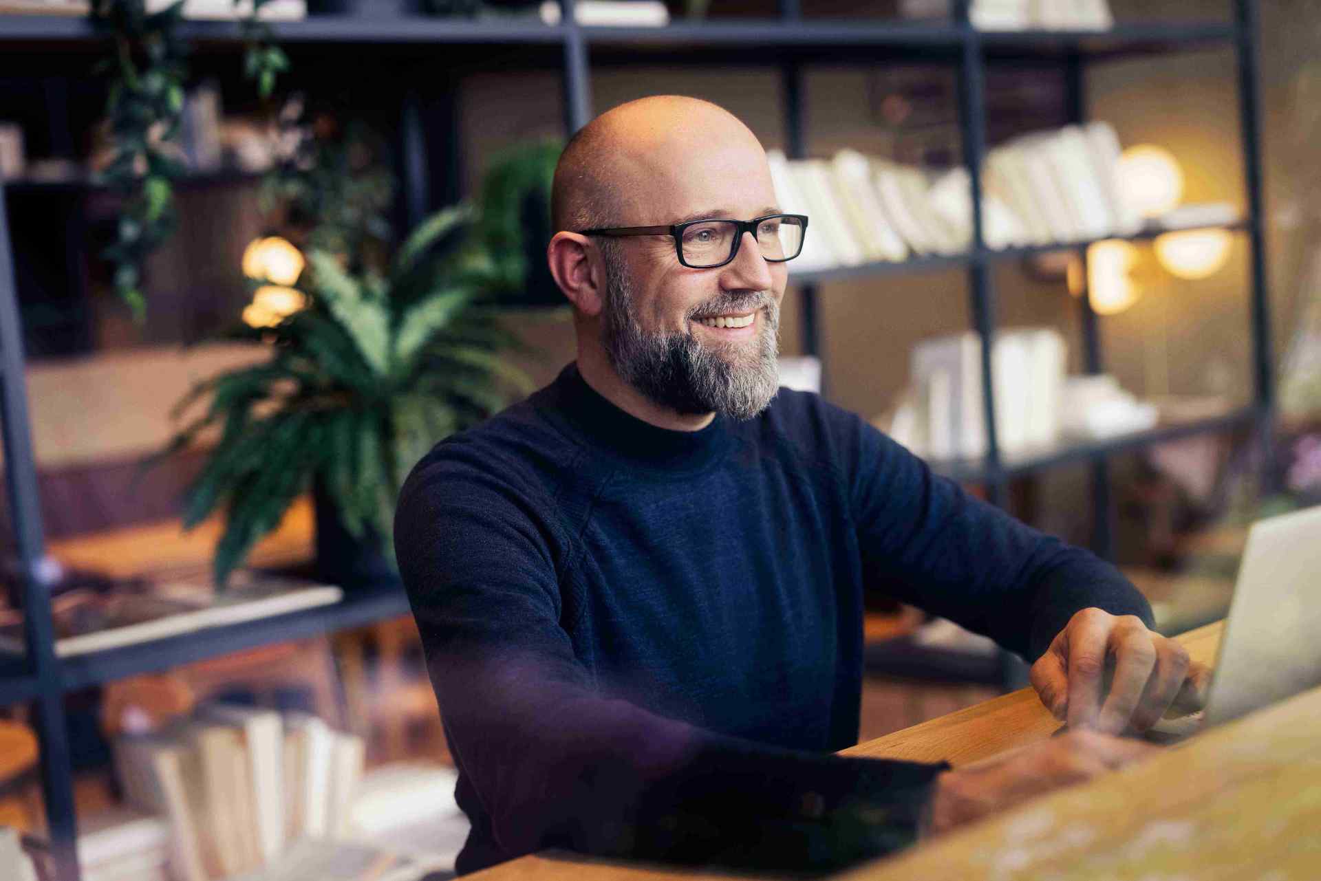 Man met bril glimlacht terwijl hij aan een laptop werkt.