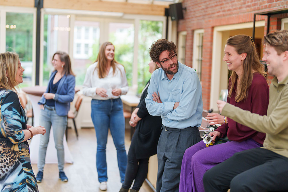 Groep mensen praat en lacht samen in een informele setting, terwijl ze in een lichte ruimte staan.