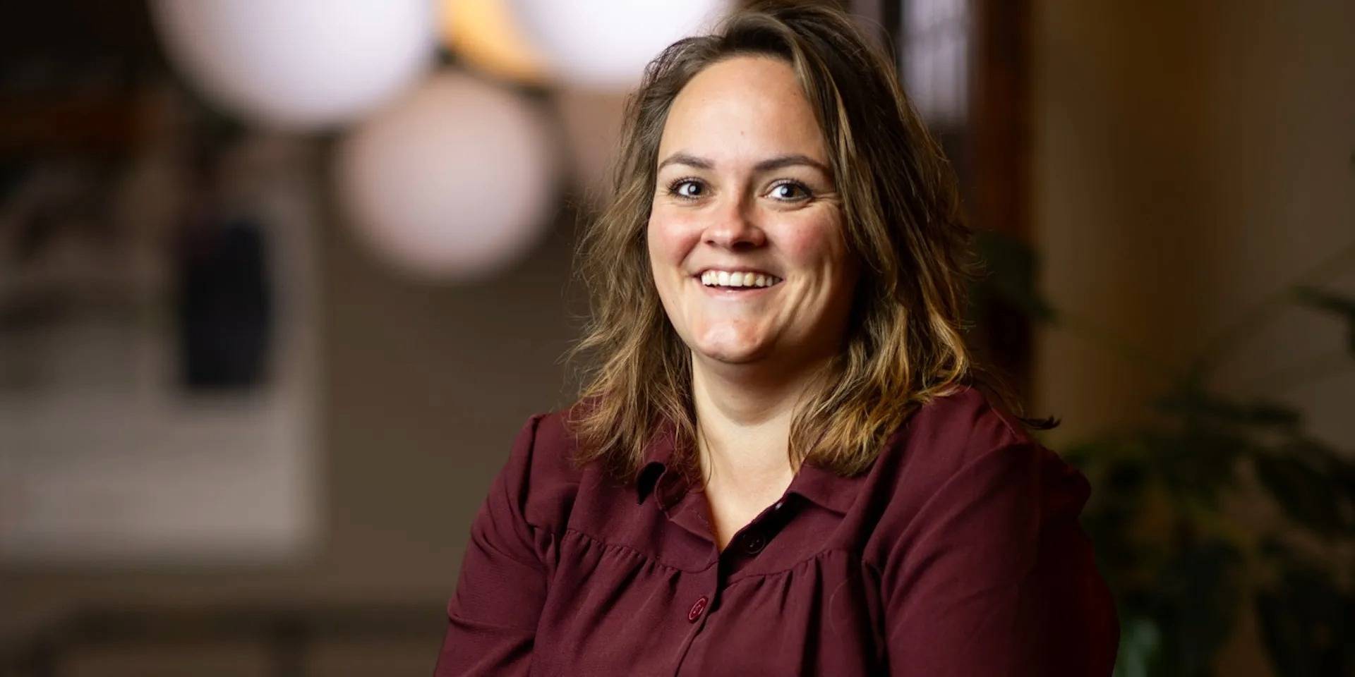 Een vrouw met halflang haar glimlacht naar de camera in een warme, sfeervolle binnenruimte.