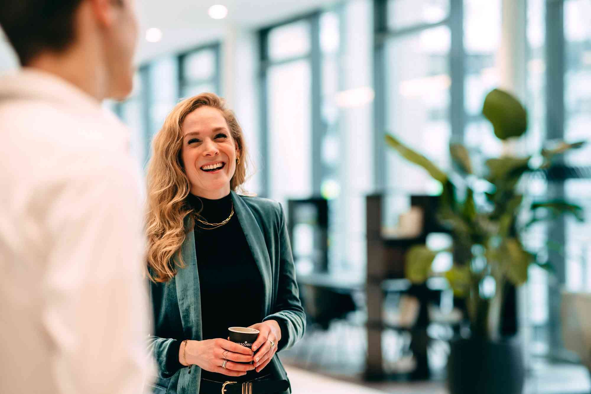 Woman laughing while talking with a cup of coffee in hand in an office.