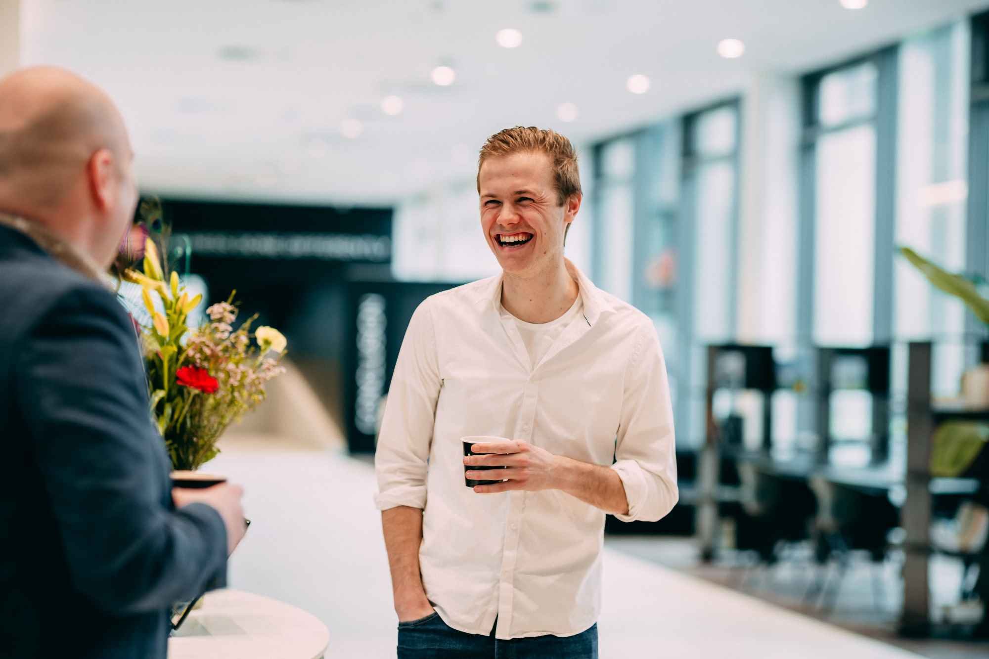 Man laughing while talking to another person with a cup of coffee in hand.