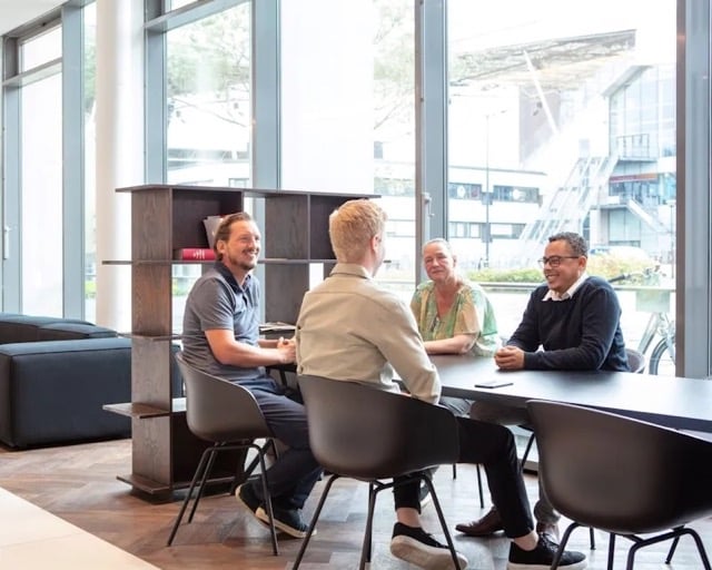 Vier collega's zitten aan een lange tafel in een openbare ruimte.