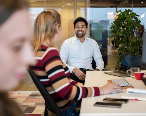 Een man zit bij zijn vrouwelijke collega aan bureau en lacht vriendelijk.