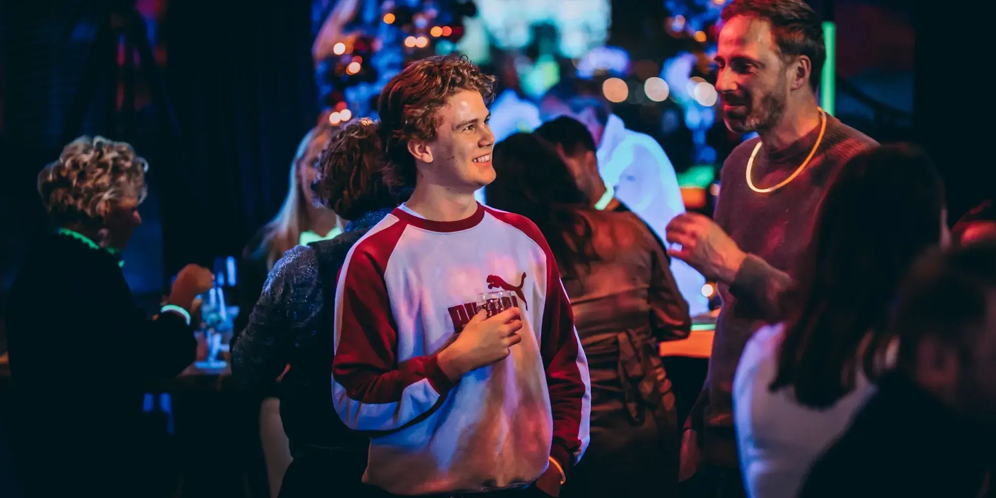 Jonge man in rood-witte trui met drankje in de hand lacht tijdens een druk bezochte borrel.
