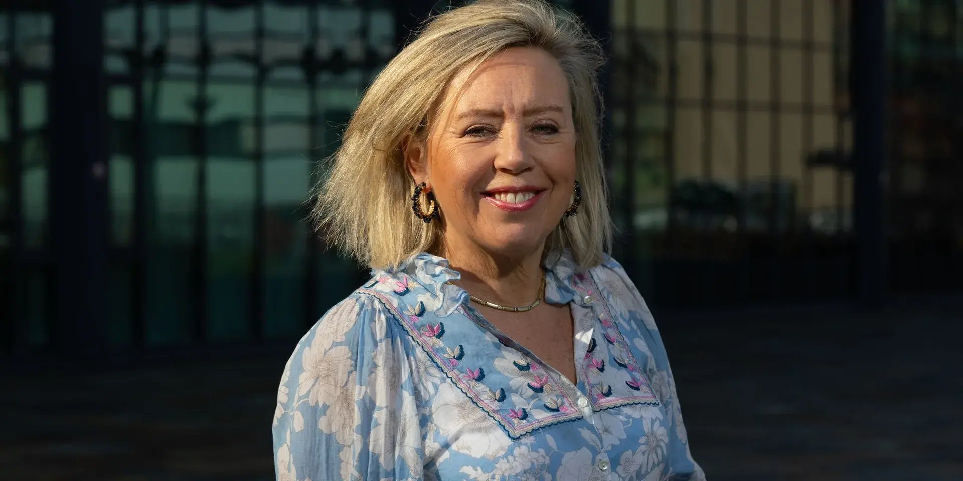 Vrouw met blond haar en blauwe blouse glimlacht buiten voor een gebouw.