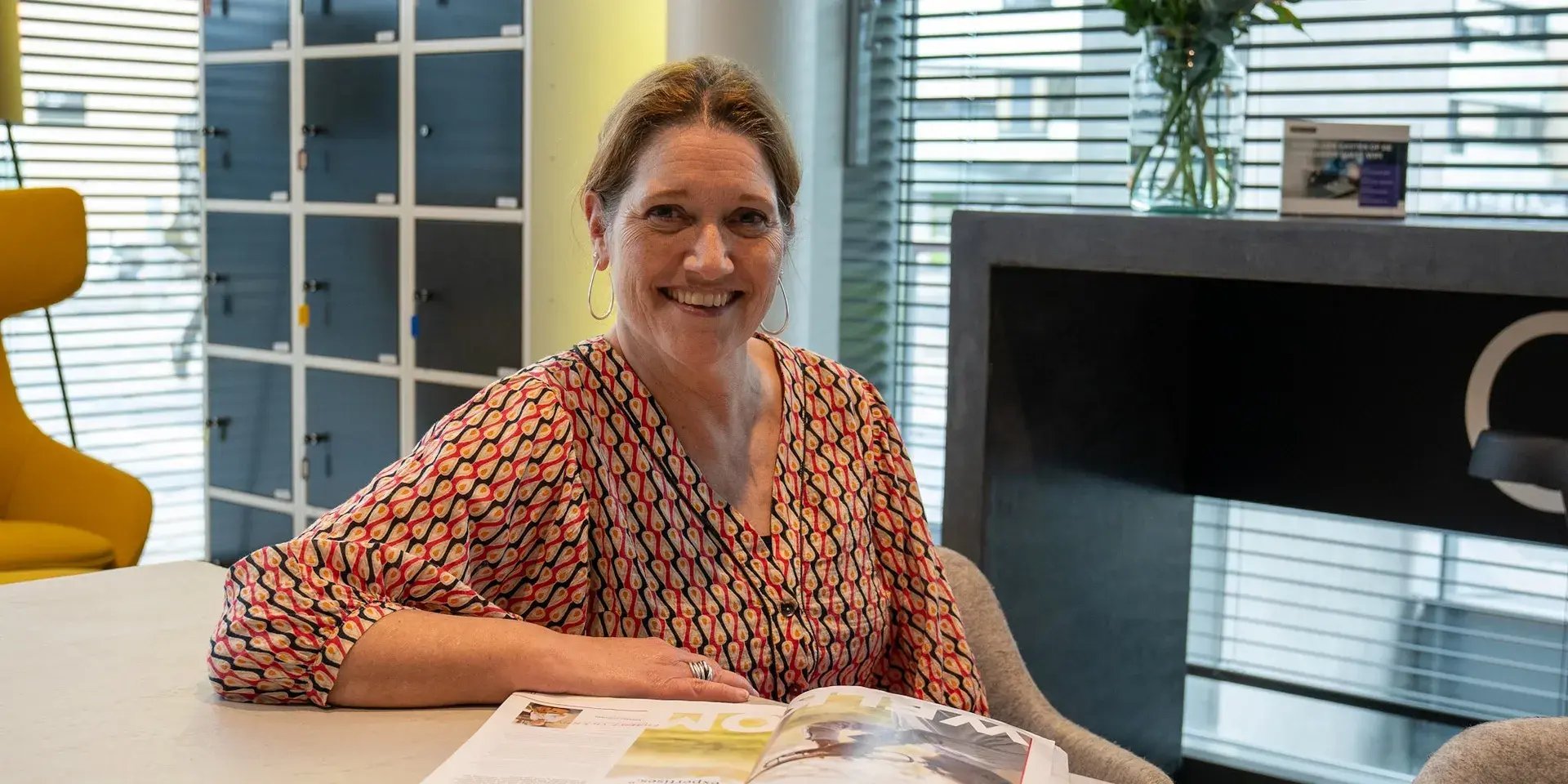 Vrouw met patterned blouse zit glimlachend aan tafel met een open tijdschrift.