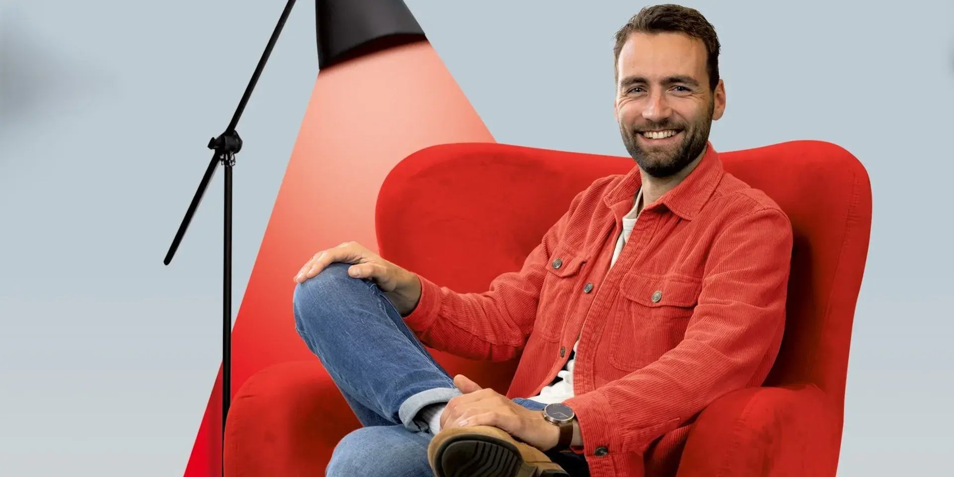 Man in rode jas zit lachend in een rode stoel met een lamp naast zich.