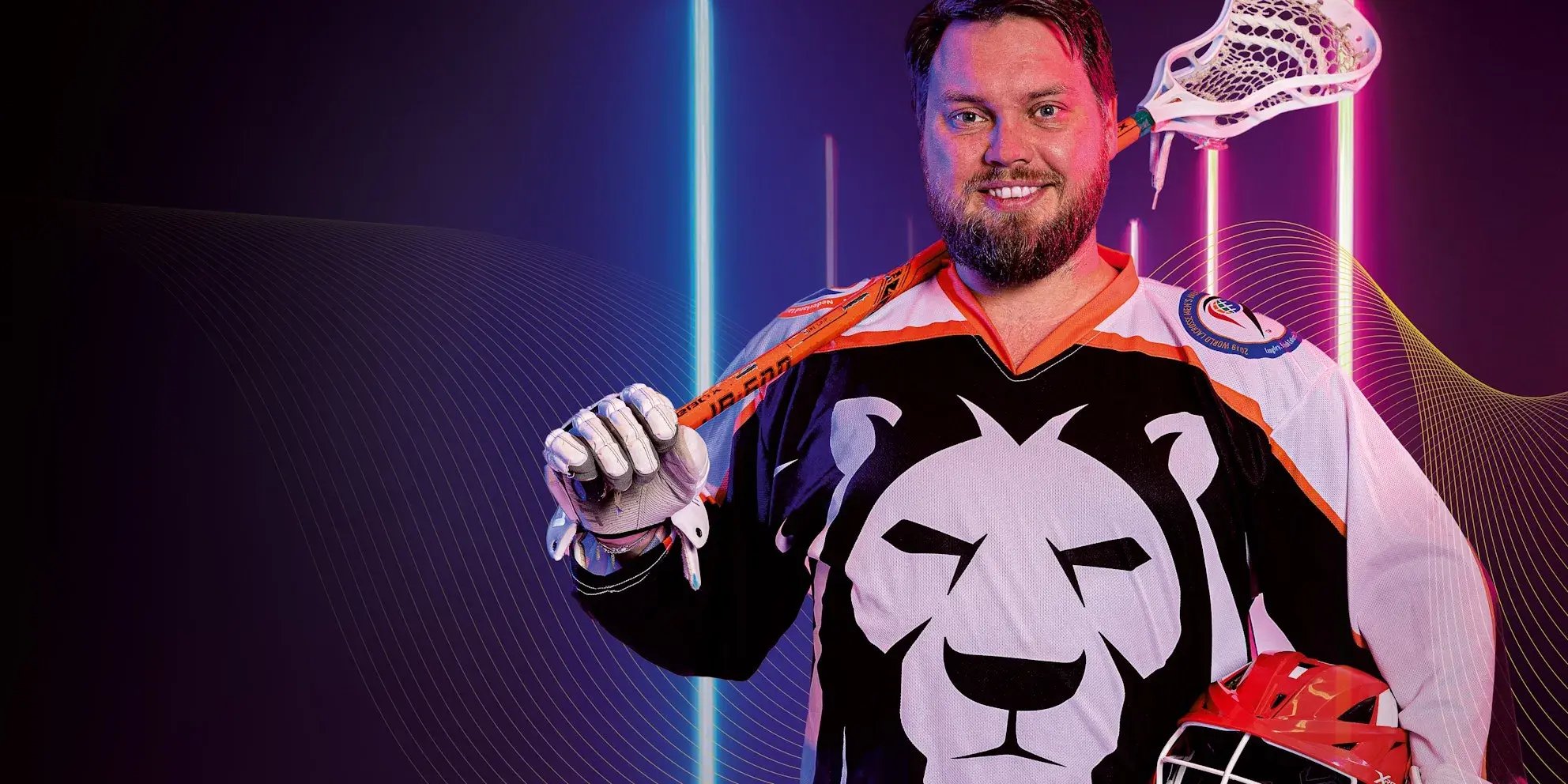 Man in sportshirt met leeuw en lacrossestick poseert glimlachend.