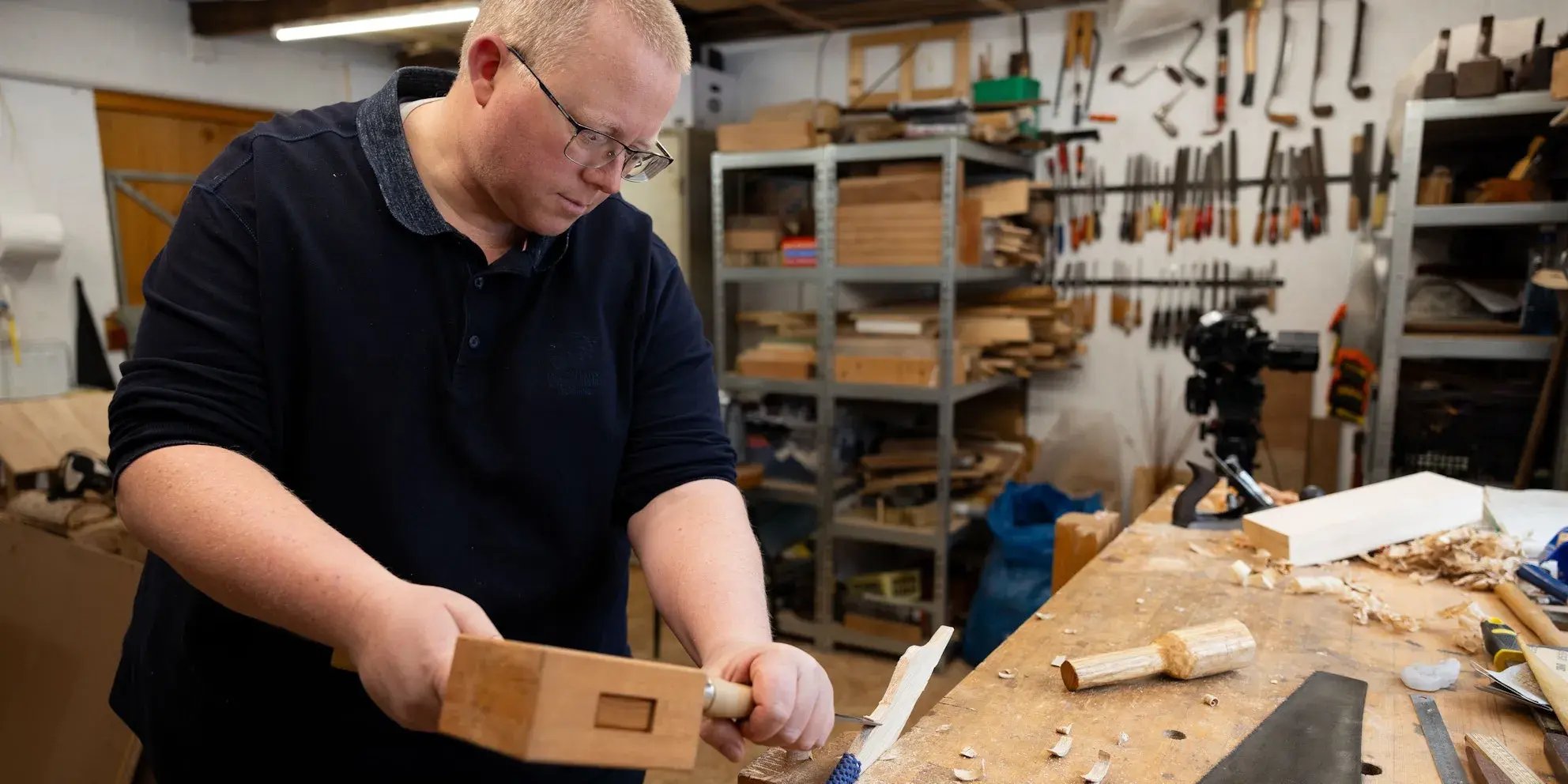 Man werkt geconcentreerd met hout en beitel in een werkplaats vol gereedschap.