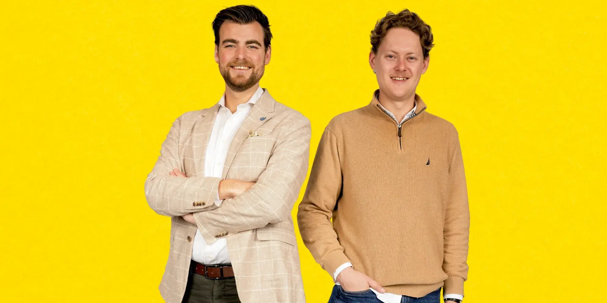 Twee mannen poseren lachend voor een gele achtergrond.