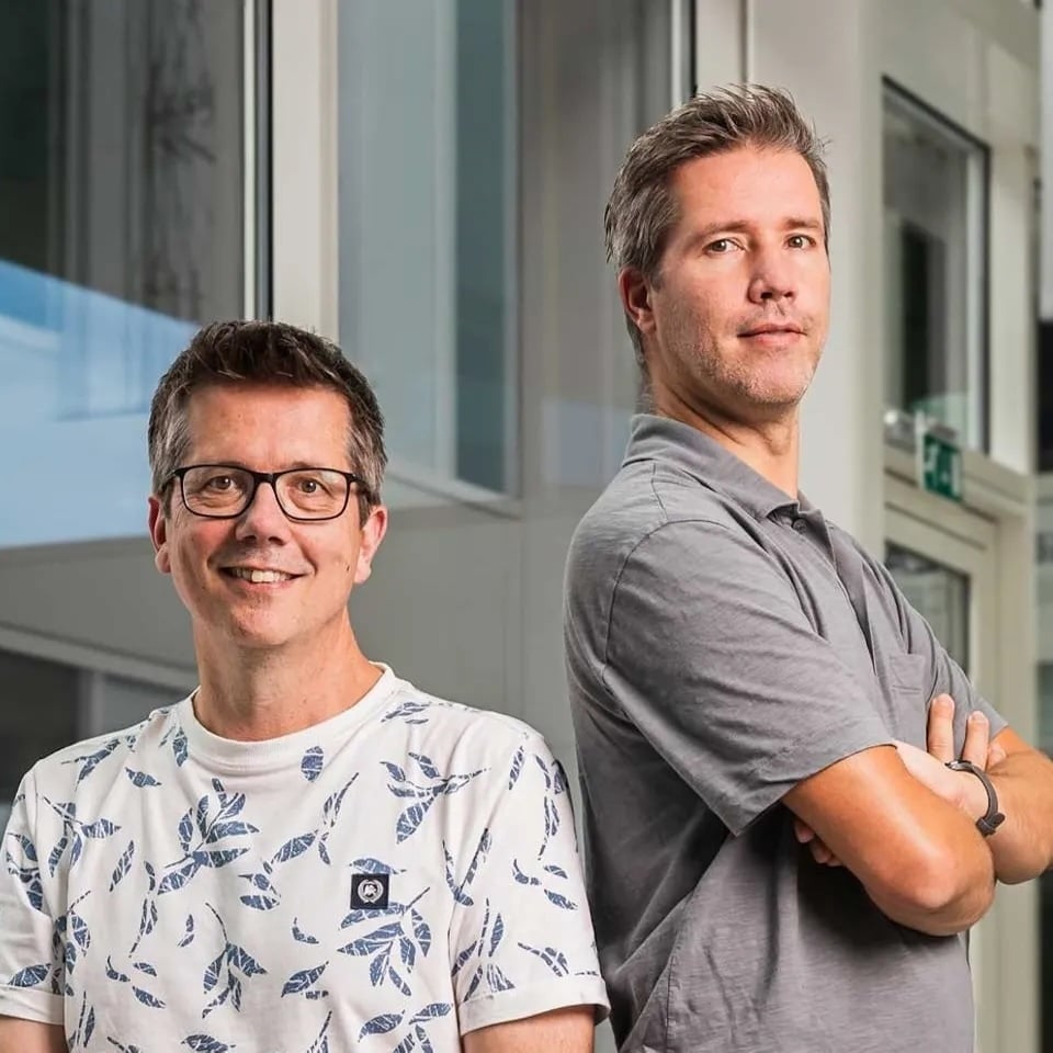 Emiel lachend in een wit shirt met Paul naast hem in een grijs shirt met zijn handen over elkaar