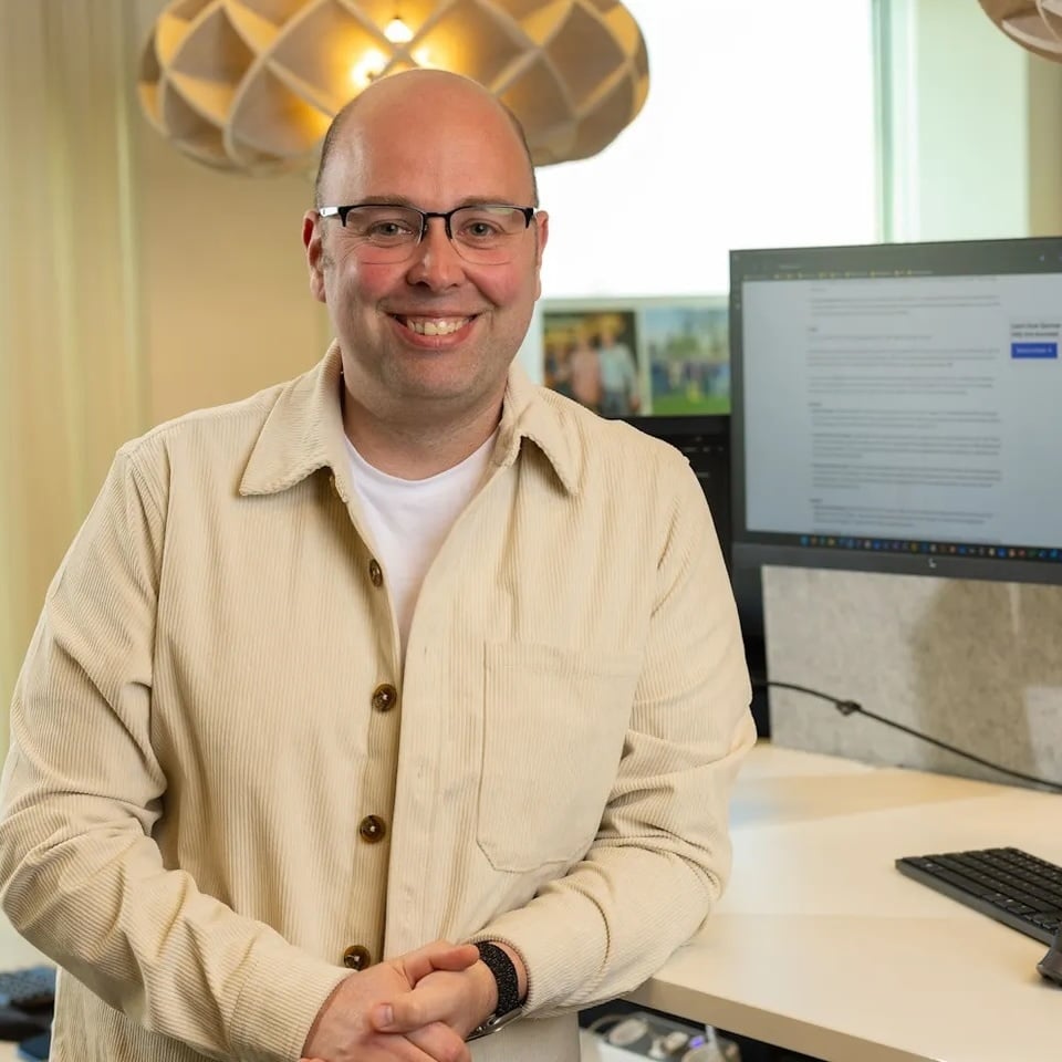 Glimlachende man met bril in een beige overhemd staat in een kantoor naast twee computerschermen.