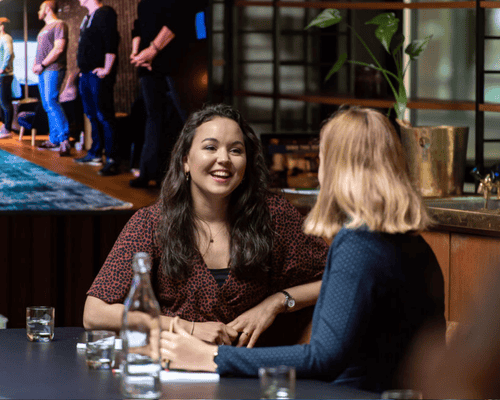 Twee vrouwen lachen met elkaar aan tafel tijdens een gesprek, met een groepsfoto op de achtergrond.