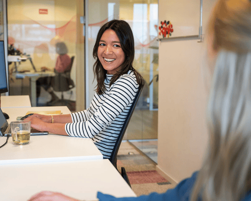 Een vrouw in een gestreepte trui glimlacht naar een collega terwijl ze aan een bureau werkt.