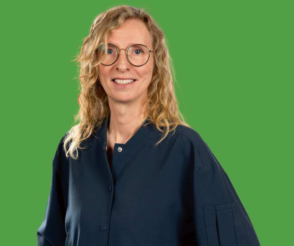 Een vrouw met blond krullend haar en een bril glimlacht voor een groene achtergrond.