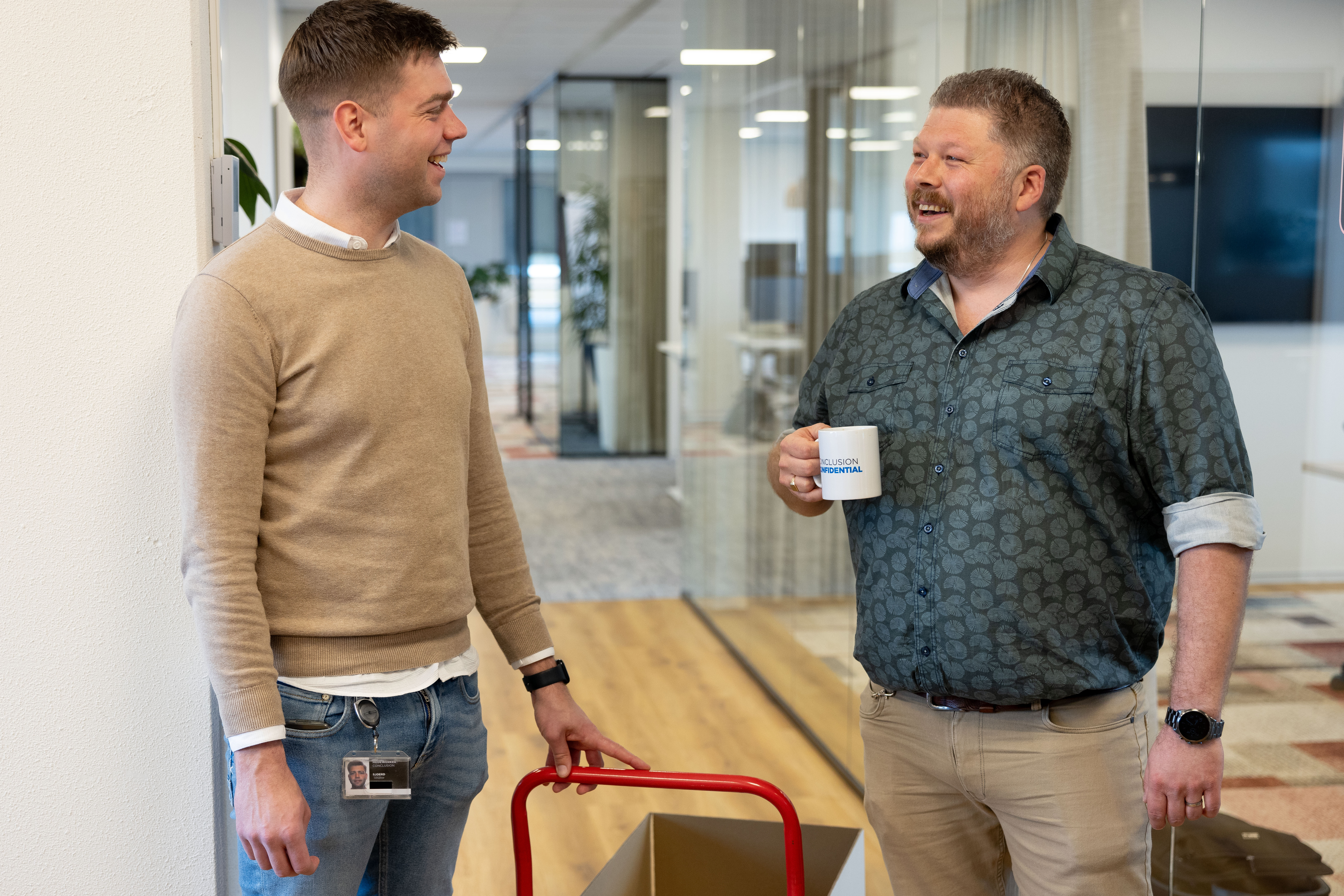 Man wearing a beige sweater and man wearing a blue shirt laughing and talking in an office space.