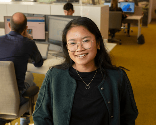 Een medewerker staat glimlachend voor een bureau, terwijl anderen aan hun computers werken op de achtergrond.