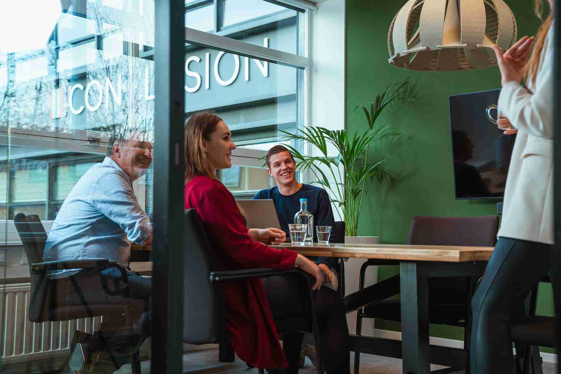 Een groep mensen zit aan een vergadertafel, lachend, in een vergaderruimte met het 